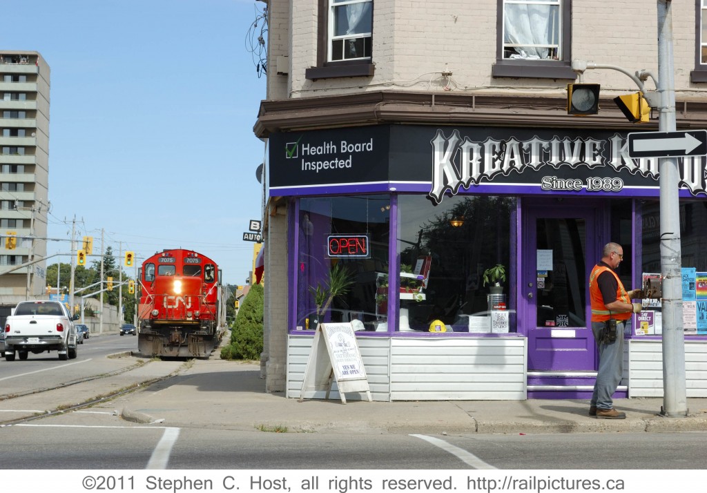 Don\'t walk: CN 580, led by CN 7075, is patiently waiting while the Conductor activates the crosswalk and traffic lights to stop opposing vehicular traffic. The CN Burford spur runs alongside Clarence St, and for a hundred feet even runs inside the street with a similar crosswalk/streetlight type system to stop traffic. Ultimately, the train obeys traffic signals and requests the right of way at least four times.