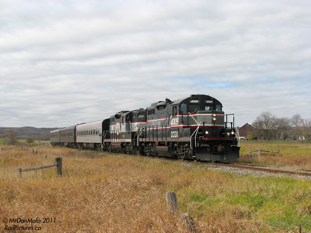Rolling through farmland with the Niagara Escarpment in the background, the Credit Valley Explorer makes it way south through Cheltemham Ontario, at Chinguacousy Road.  The unusual doubleheader was the official \"Farewell 1000\" trip as CCGX 4009 had recently arrived to replace it. The somewhat tired 1000 continued in use for a few more months playing second fiddle, until it was finally shipped out to the Lafarge plant in Bath ON for local switching. The rebuilt 4009 became the Orangeville-Brampton Railway\'s main and only power.