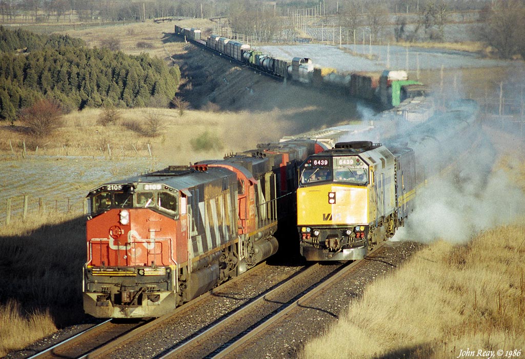 A westbound VIA train has just come to a stop in a cloud of steam after being nailed by the hotbox detector at Newtonville in March 1986.  CN 393, led by HR-616 2105 and two six-axle MLWs passes the VIA train\'s head-end.