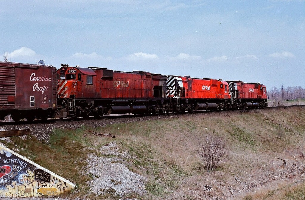 Departing Agincourt Yard May 4, 1980, CP Rail train #942 enters the high rail at Staines, Ontario with a seventies superpower trio 4729 (M636) / 4565  (M630) / 4737 (M636). The big M units had a propensity to leak – anything – witness the fresh oil stains on 4729\'s front fuel tank, 4565 is in fresh Multi Mark  paint and shows evidence (blistering paint) of possible exhaust leak and/or turbo overheating below the exhaust stack and likely the head end crew is happy they are not occupying leak prone 4737 where the cab is covered in oil thrown off the sixteen cylinder ALCO designed engine. Note the CP script on the fifty foot box car behind 4737 and the graffiti is on the Old Finch Ave. underpass. Kodak Kodacolor II ASA100  negative transported by a Nikon Nikkormat EL. Photographer S. Danko.