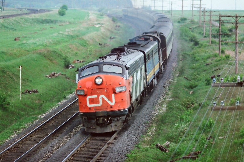 At track speed: a twelve car train powered by a FP-9A-FPB4-RS18 (the latter a 3100) lashup is Via CN train #63 the Rapido  at mile 280 CN Kingston subdivision on the approach to the Clarke crossovers in this spring 1977 negative. Note the jointed rail and even so track speed on this super-elevated curve is 90 m.p.h. ( Turbo Trains restricted to 95 m.p.h. (154 k.p.h. in metric-eze)). The late 1970\'s transition from CN & CP owned/operated passenger service  to Via Rail provided many interesting  power & equipment combinations. Photograph location is the ever popular Stephenson Road/Lakeshore Road wooden bridge also referred to as Lovekin – the former station site on the adjacent CP Belleville sub. Kodak Kodacolor II ASA100  negative transported by a Nikon Nikkormat EL. Photographer S. Danko.