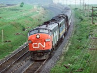 At track speed: a twelve car train powered by a FP-9A-FPB4-RS18 (the latter a 3100) lashup is Via CN train #63 the Rapido at mile 280 CN Kingston subdivision on the approach to the Clarke crossovers in this spring 1977 negative. Note the jointed rail and even so track speed on this super-elevated curve is 90 m.p.h. ( Turbo Trains restricted to 95 m.p.h. (154 k.p.h. in metric-eze)). The late 1970's transition from CN & CP owned/operated passenger service  to Via Rail provided many interesting  power & equipment combinations. Photograph location is the ever popular Stephenson Road/Lakeshore Road wooden bridge also referred to as Lovekin – the former station site on the adjacent CP Belleville sub. Kodak Kodacolor II ASA100  negative transported by a Nikon Nikkormat EL. Photographer S. Danko.