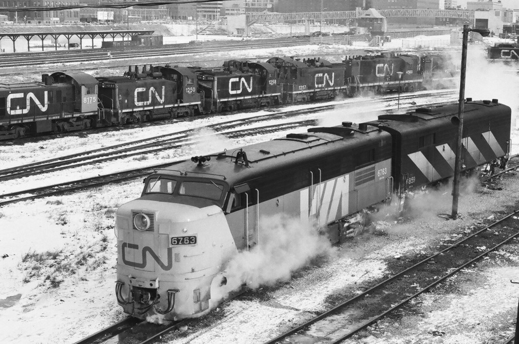 Cab Units: FPA-4 Via CN 6763 and FPB-2u CN 6859 at the CN Spadina motive power shop, February 1977. 6763 is in the short lived \'Via CN\' paint scheme complete a red CN noodle on the yellow nose. 6859 is one of only two CN FPB-2u units. Note the eclectic mix of switchers in the background. Front Street is visible to the left behind the freight loading dock and  behind the TTR  John Street Tower (complete with a full roof – today it is chopped off)  is the newer pedestrian bridge to the new CN tower. Kodak Plus X Pan b&w negative film, ASA125, transported by a Nikkormat. Photographer S.Danko.