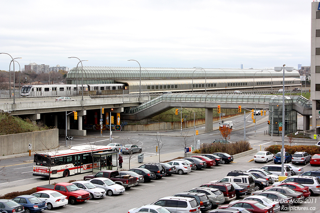 A new Toronto Rocket trainset pulls into the 1970\'s era Yorkdale Subway Station, as a 47B Lansdowne bus waits for passengers nearby. On the right, passengers travel to and from the subway via the matching passage connecting to the sprawling Yorkdale Mall.