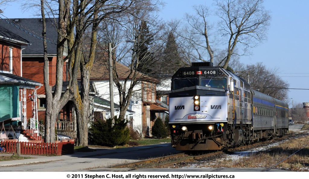 Would you live on this street? Train #85 with VIA Rail 6408, painted in Coors Light livery is heading alongside the \"street running\" of Guelph. You can see the homes and porches along the north side of Kent. St, and the south side of Kent is to the right of the picture, also with homes facing the street and railway. Kent St. is very close to downtown Guelph and pre-dates the arrival of the Toronto and Guelph/Grand Trunk Railway in 1855.