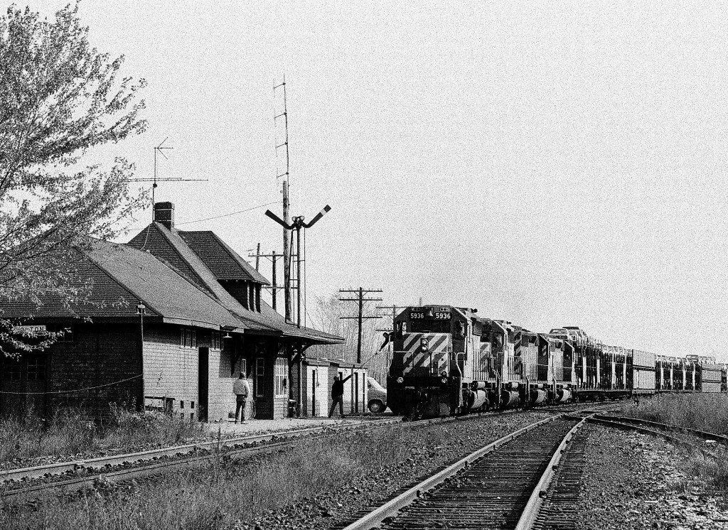 The hogger at the controls of CP Rail train #415 (#5936 ) receives the train orders on the fly from the CP Rail Bolton Ontario station operator. The quartet of SD40-2\'s is the standard power for CP train #415  the Pacific Auto Train (PAT) which at that time was THE hot shot freight to the west coast. Note the open bi-level and tri-level auto carriers and CP still had  jointed rail at this location. October 22, 1983 Kodak Plus X Pan b&w negative film, ASA125, transported by a Nikon Nikkormat FT. Photographer S.Danko.