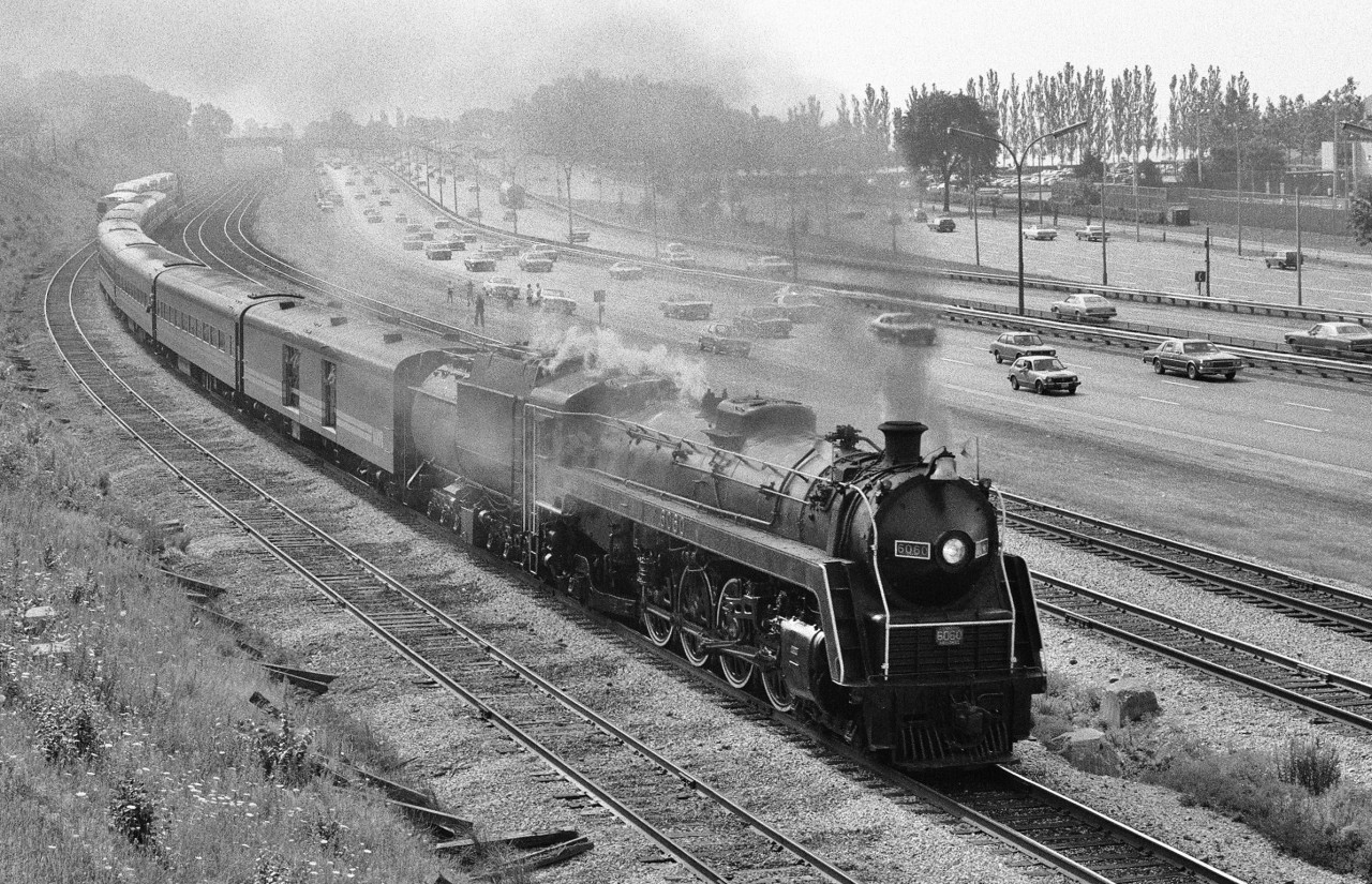 Sharp staccato stack talk wakes the Saturday morning Sunnyside Ontario neighbourhood as the  westbound expressway parade paces MLW built CNR Mountain type 6060 on August 23, 1980. The Upper Canada Railway Society car 13 Cape Race holds the markers on this train. At extreme left note the single level 100 series GO control cab car leading the approaching GO train on track 1 CN Oakville Sub. Perhaps viewers with detail knowledge of this train will be willing to share. Kodak Tri X pan  b & w  ASA 400 negative. Photographer S. Danko.