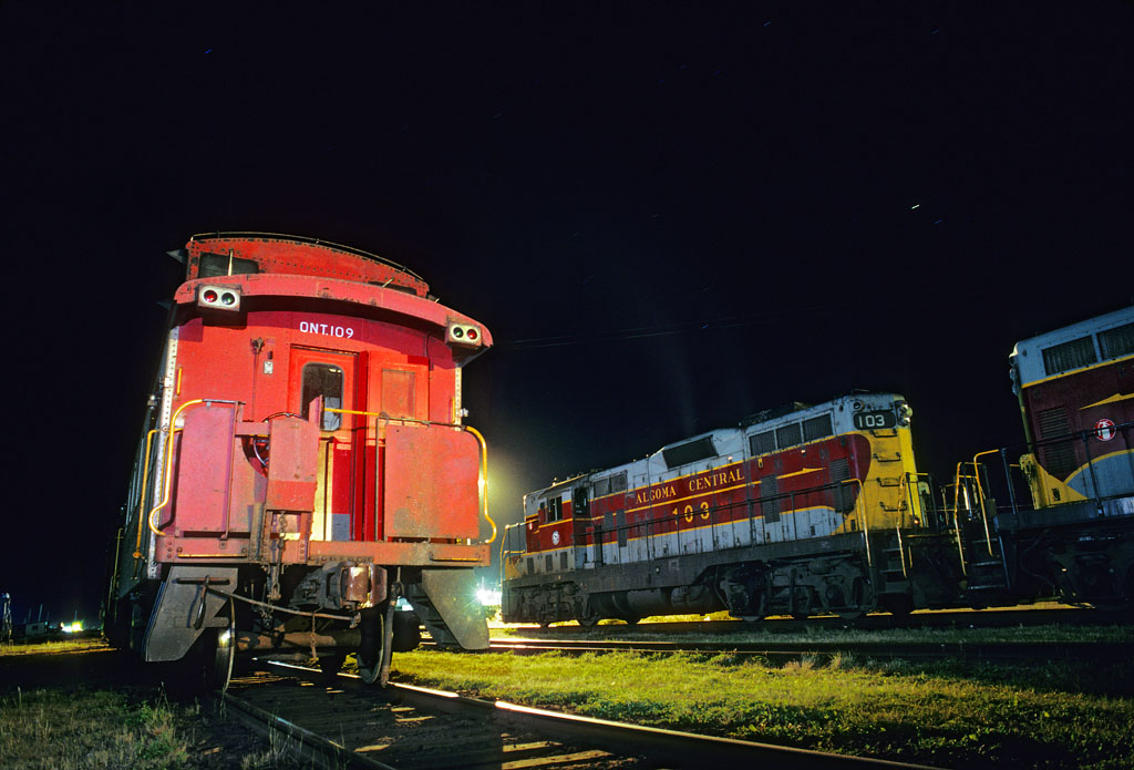 A pair of Algoma Central GP7s rest alongside an Ontario Northland caboose at Hearst, Ontario, on the night of September 9, 1993.