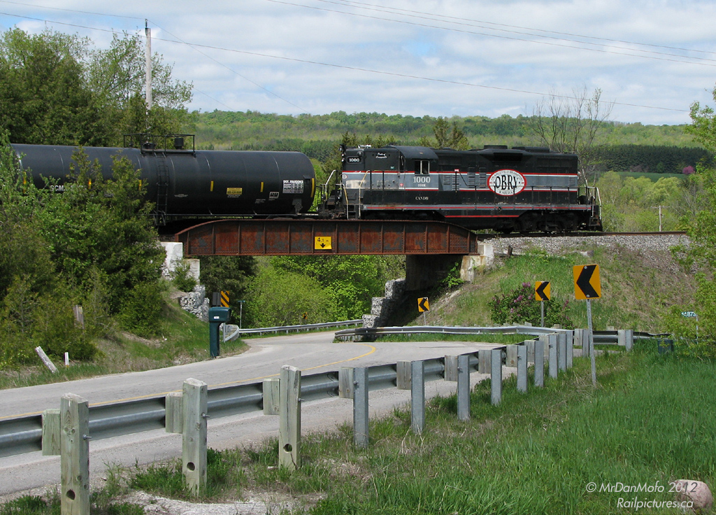 With the rolling hills of the Niagara Escarpment coming into view in the distance, CCGX GP9 1000, the sole power for the Orangeville-Brampton Railway shortline, heads north to Orangeville with a few cars from CP, crossing the bridge at Ferndale over the twisting Chinguacousy Road.