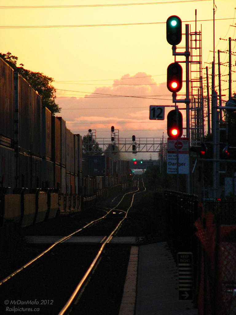 CN intermodal #149 climbs out of downtown Brampton on the ascent to Mount Pleasant, with clear signals all the way to the Credit River. In the foreground, another \"high green\" beckons an unknown freight on the north track which will come howing through close on #149\'s tail, chasing the fading orange glow of another day\'s passing.