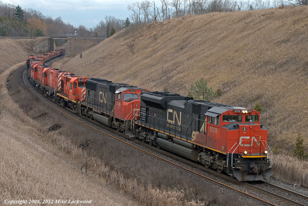 CN 8009 and 5639 lead six retired MLW's on CN 305 at Beare. On their way to Chicago are NBEC 1840, SFEX 4203, NBEC 4210, SFEX 4202, SFEX 4204, and NBEC 4214, all of CP heritage and all framiliar with this location, albeit crossing the bridge on the Belleville Sub in the background. 1353hrs.
