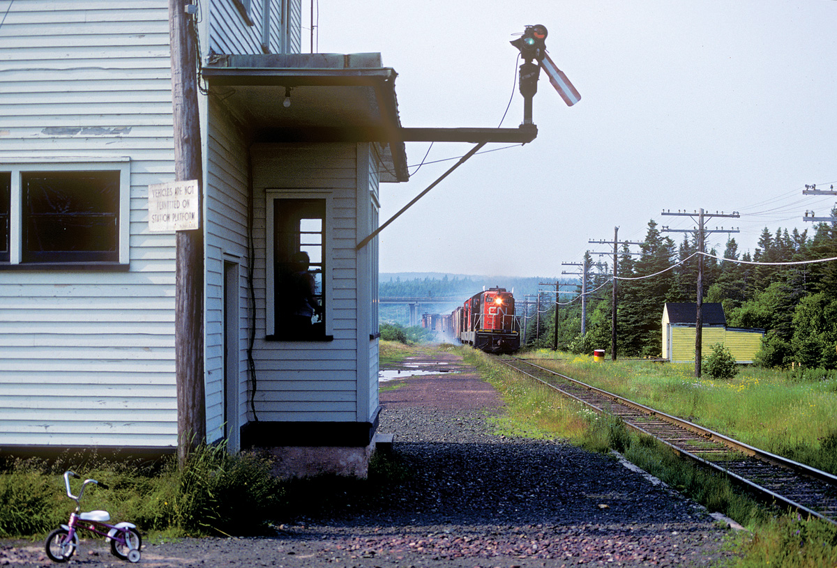 Remarks: Train 204, the mainline freight from Port-aux-Basques (ferry terminal and yard on the southwest coast) to St. John's, approaches Goobies in a cloud of brake shoe smoke (there were no dynamic brakes in Newfoundland). Note the agent looking out the window at the arriving train. Goobies was on CN's St. John's Subdivision, 20.9 miles east of Clarenville.