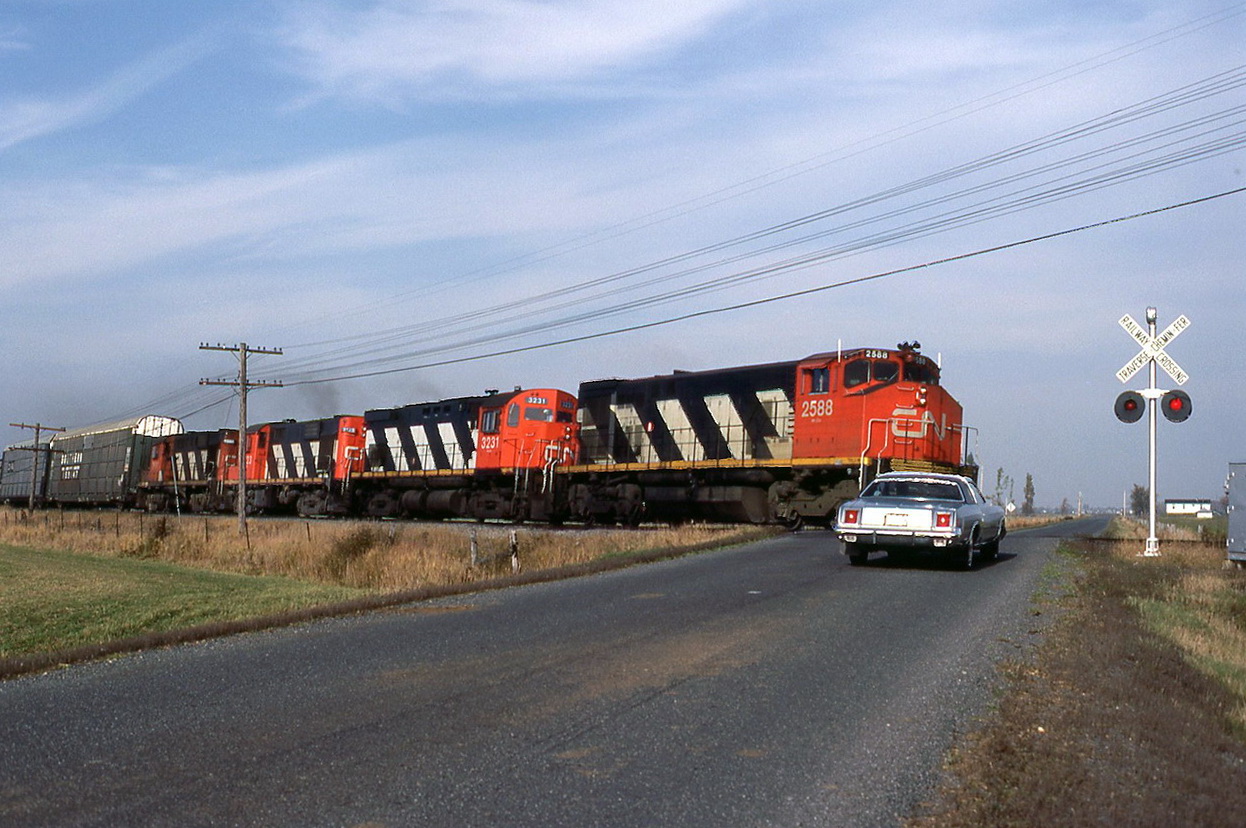 CN 345 with 4 MLWs,the popular power of those days passes the crossing at a good speed but the Dodge got there a split second before.