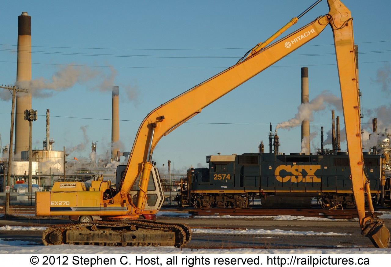 CSXT GP38-2 2574 is shown switching the north end of the Sarnia Yard framed between  the Boom/Stick of a Hitachi EX270 excavator. Behind the locomotive are the smokestacks of the omnipresent Imperial Oil Sarnia plant. This plant along with the other large petrochemical concerns in the area provide lifeblood to the CSX in Sarnia despite almost 20 years of relative isolation from the rest of the network.