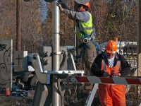 A signal maintainer goes about his business finishing up the gate wiring for this newly installed crossing signal, meanwhile his apprentice mounts a bracket to the mast before mounting the sign to it depicting how many tracks this grade crossing has. 