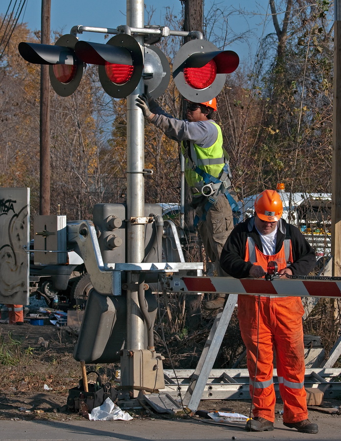A signal maintainer goes about his business finishing up the gate wiring for this newly installed crossing signal, meanwhile his apprentice mounts a bracket to the mast before mounting the sign to it depicting how many tracks this grade crossing has.