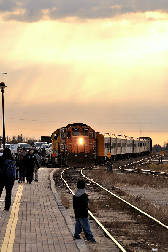 Playing in the sand at the Cochrane station, left from years of engines struggling to grip slick, slippery and icy rails during departure from the Cochrane station. Exploring the world he's in and enjoying the small beach created between the platform and rails a rumble approaches to the east. He looks up. A roar from Ontario Northland GP38-2's 1806 and 1809 as the Engineer increases the throttle to pull 4 flatbeads loaded with vehicles out of the ramp track and onto the main. The bell starts ringing to warn the awaiting passengers on the platform of the train's approaching presence. Startled, the boy in the picture runs to his parents side. In minutes the train will pull up to the station and into position to start loading the almost 300 people standing around to board for Moosonee. Thanksgiving Monday is a busy day for the northbound Polar Bear Express.