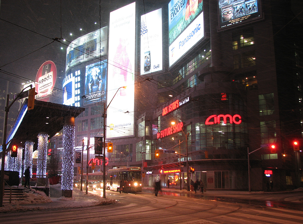 It's a snowy and frigid winters' night at Dundas Square. Billboards advertising mobile phones, movies, the State of PA, and local stores light up the night sky as a stopped streetcar loads passengers in the cold. Just another Toronto night downtown.