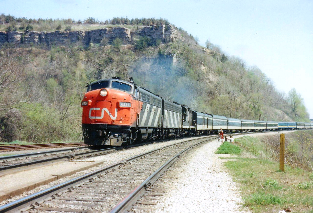 A glorious spring day at Dundas, a young mother and children have just climbed the hill by way of the Grindstone Creek viaduct and are waiting for this westbound CN passenger to pass. Power is 6534, 6623 and 4103.
Despite it being only noonish, it is a very warm day. Direct center top is Dundas Peak where some great views of CN action can be noted.