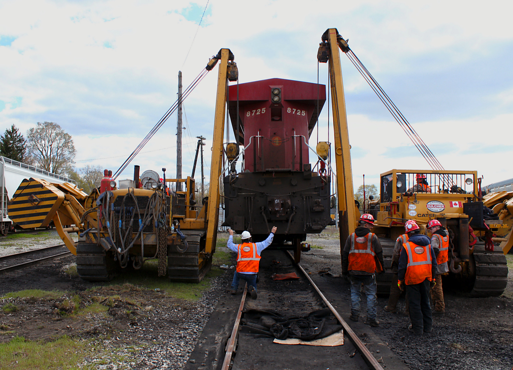 CP 8725 is skillfully "conducted" back down onto it's new dummy axle by the Hulcher foreman. Other members of the Hulcher team watch, taking a well deserved break after wrestling the defective wheel set out. In total it took an hour and a half to complete the job. Photo taken with permission by a CP Mechanical Services employee wearing all the required PPE.