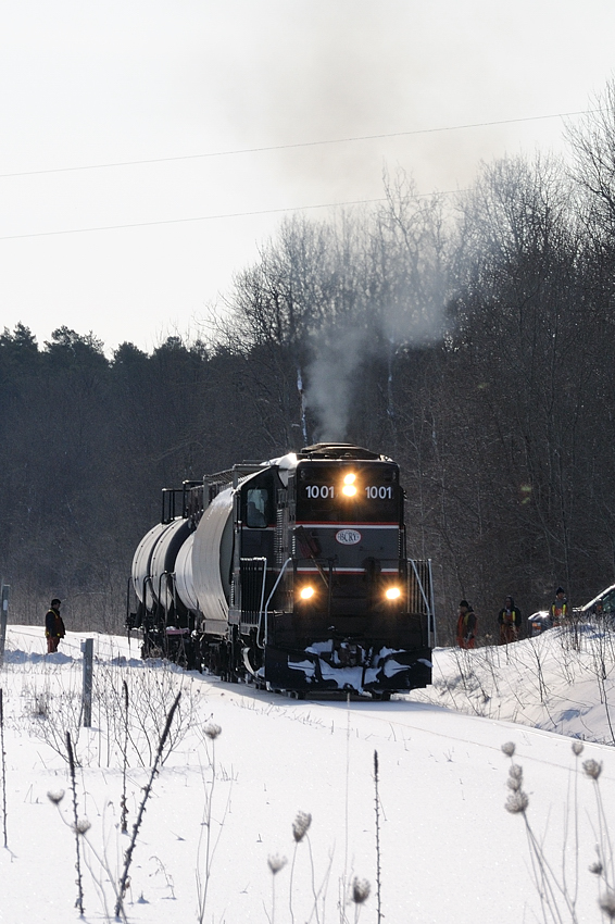Barrie-Collingwood Railway chase 1 of 7. In February 2009 I set out on a mission to capture the infrequent train to Collingwood, Ontario. Arriving at the BCRY shops around sunrise I talked with some employees to confirm the train would be running to Collingwood. Sometime after 08:00 the crew brought the engine out the stall and began their minor switching duties grabbing three cars destined for Canadian Mist distillers at the end of track in Collingwood. After waiting for a northbound CP the crew was given the light and proceeded westbound. A few employees followed the train westbound, protecting the grade crossings and chipping out any build up of ice in the flanges of the crossings. In this scene those employees watch as the engineer throttles up the old GP9 as their westbound train crosses the Sunnidale Tosorontio Townline along County Rd 10, just west of Angus, Ontario.