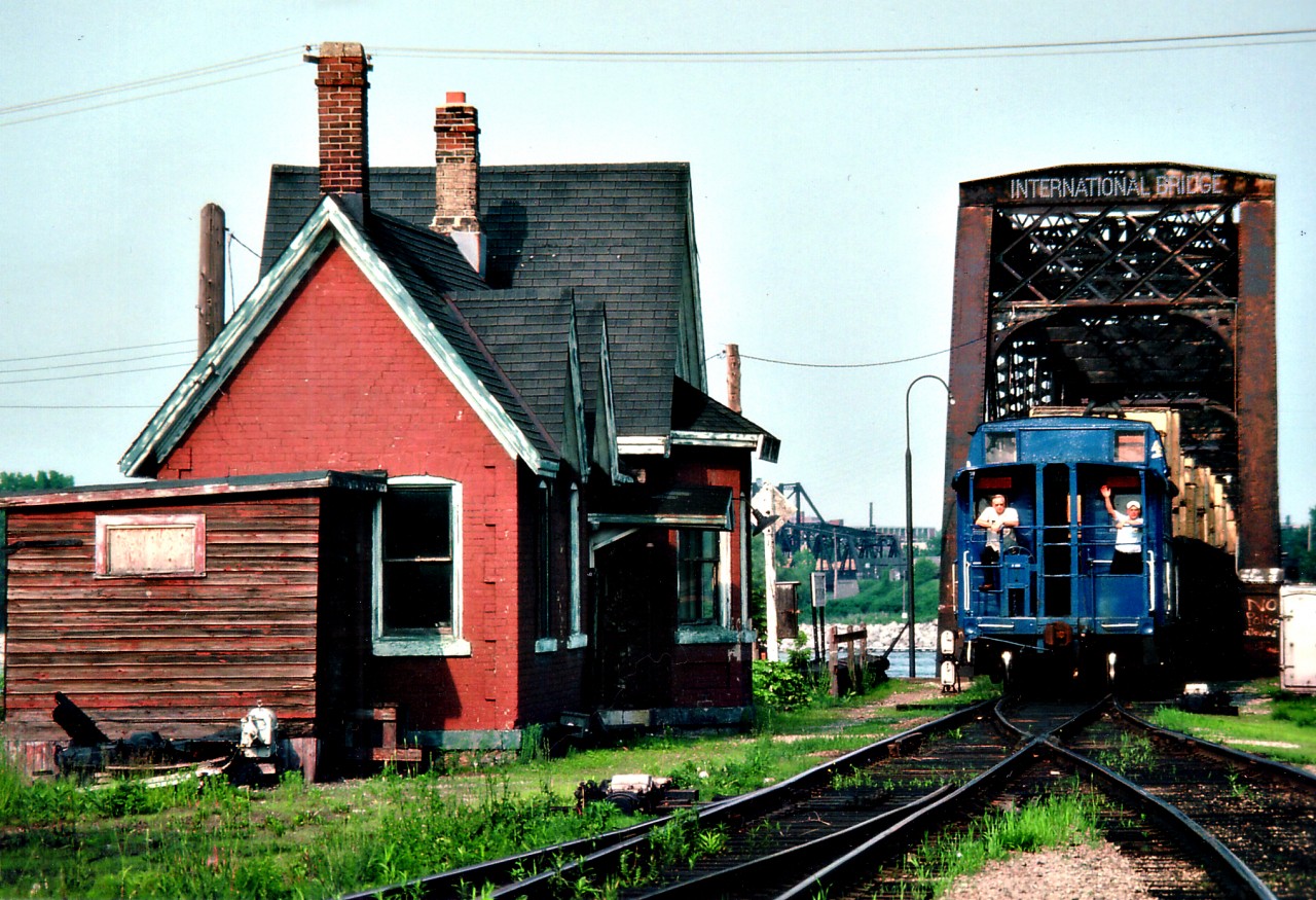 Like a scene from long-ago branchline railroading, Conrail crew watches and waves from the Van as the CR transfer heads back Stateside on a late afternoon in June of 1979. Note the US-bound merchandise is in crates. The vans are history, as is the old Bridgeburg B-1 Station. The building was jacked up and moved across the tracks to be trucked to the RR theme park on Central Av to keep CN 6218 and Ridgeway Sta company. Why it was then painted a sickly yellow I don't know. Bridgeburg was the original community name. Fort Erie was further to the south. Now, of course, they are one.