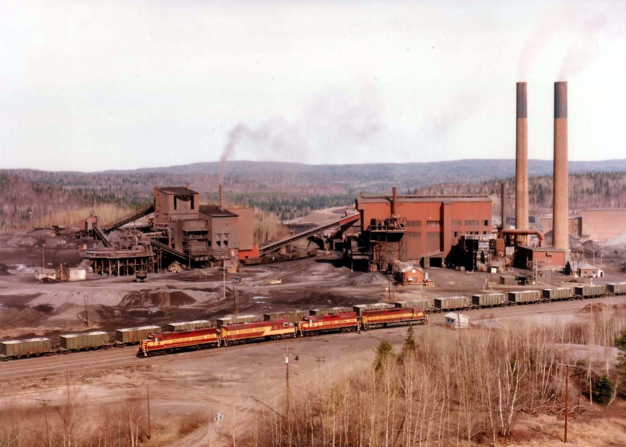 From high on the rocks, here is an overview of the great Sinter Plant that used to provide so much employment to the town of Wawa, Ontario as well as the former Algoma Central Railroad. The purpose of the plant was to supply blast furnaces with sinter, a combination of blended ores, fluxes and coke which is partially "cooked" or sintered. In this form the materials combine efficiently in the blast furnace and allow for more consistent and controllable iron manufacture. From here, material was transported out to Michipicoten Harbour, about 12 miles west, for GreatLakes shipping. Awaiting next assignment are WC 6604, 6655, 3012 and 6506. The F45, 6655, was retired in 2002. Later on this year 1998 the plant was closed, buildings removed, track removed, and nothing is left but a blight on the landscape.  The plant had been in continuous operation since 1939, and is now but a memory.