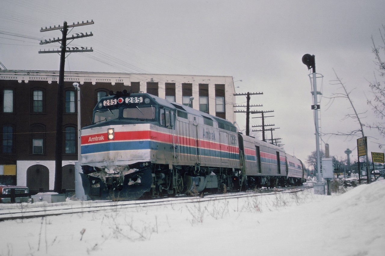 Amtrak 285 leads "The Rainbow" toward the steel arch bridge and the USA in this view from January 28, 1979. The train is seen crossing Queen St. in Niagara Falls. Background building is the King Edward Hotel, also now a part of history. On the right in the far background one can see a revolving tower down in the tourist district.