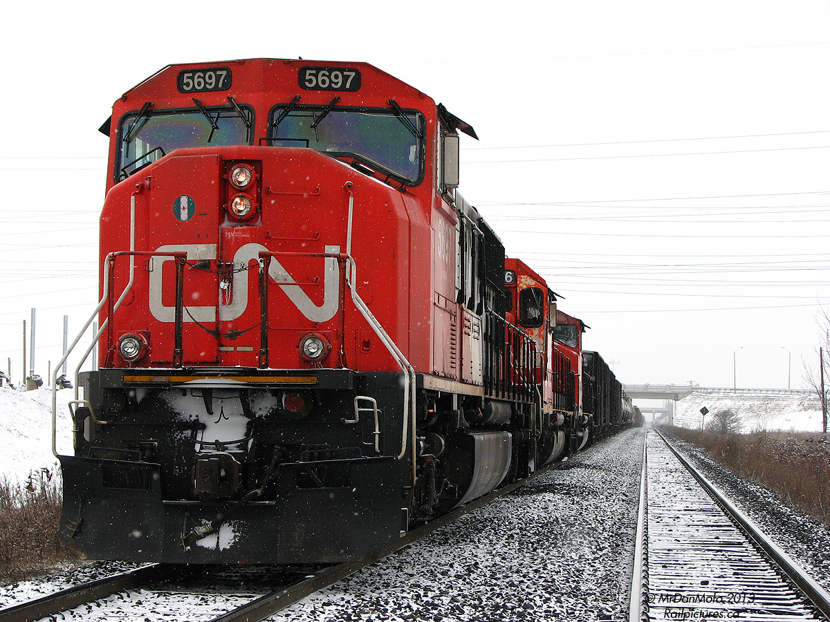 On a cold afternoon in January, CN 338 awaits a new crew at the now-closed Chinguacousy Road crossing. CN SD75I 5697, and SD40-2/W units 5386 and 5249 idle on the south main track around Mile 17 of the Halton Sub, as the snow lightly falls around them as far as the eye can see.With a newly constructed overpass of fresh white concrete behind the photographer, a transformer station to the right, a new neighbourhood under development to the left, and an old decommissioned grade crossing front, it made for an interesting area at the edge of development in Brampton, as the city limits pushed outwards and countryside that was once next to the rail lines became swallowed up over time, like snow enveloping the barren landscape in winter.