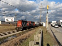 <i><b>Trucks, Trains and Traffic</b></i>. <br><br>Rolling past traffic on the busy Steeles Avenue, two GP9RM's lead CN #559 back to the mainline along the Torbram Industrial Spur through industrial Bramalea, having just finished their afternoon switching duties. The spur runs east from the Halton Sub mainline along Steeles Avenue, to get to the industrial area in the east end of Brampton near Torbram and Queen Streets. While Steeles is often choked with tractor trailers serving the manufacturing, fabrication and warehousing facilities along southern Brampton, suburban traffic congestion and stoplights are of no concern to this train.