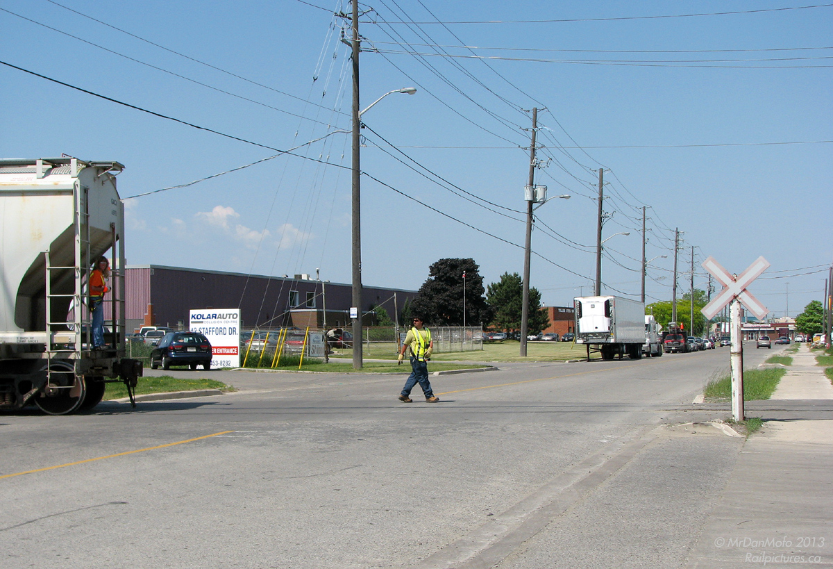 Backing down the Peel Village Industrial Lead with a cut of hopper cars for Canada Colours and Chemicals, CN local 559 slows down to cross Stafford Drive. The conductoress rides the tail car of the cut being shoved down, while her conductor trainee protects the crossing (stops any auto and pedestrian traffic) for safe passage.  Populated by commercial enterprises, autobody repair shops, fabrication and manufacturing plants, and moderate industry (once including the old AMC Brampton automotive assembly plant), the area bordered by Kennedy, Queen, Steeles and Hwy 410 near Peel Village was the traditional Brampton manufacturing base before industry started appearing around Steeles Ave in Bramalea and in the east part of town.