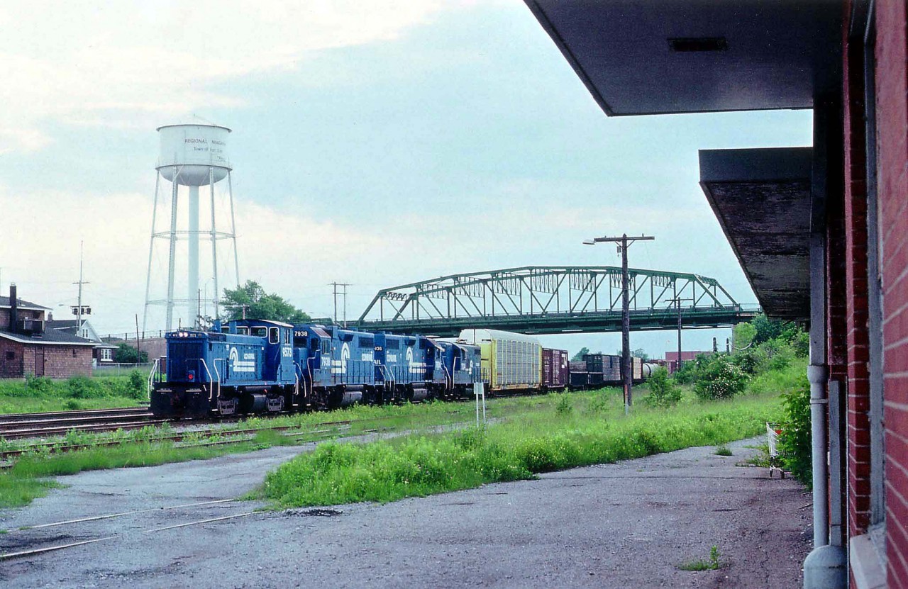 Keeping dry and out of a minor summer rain by standing under the canopy of the old Bridgeburg flat roof passenger station at Fort Erie provided a not too common angle for catching a late afternoon Conrail transfer. It is seen heading Stateside, CR 9379, 7938, 7936 and 7524 with a rather long train have just passed under the Central Av bridge. On the extreme left is the old CN crew hostel, now gone, as is the water tower, a lot of the track, and the station.  Bridgeburg was the name of the community that sprung up at the International Bridge rail crossing, it was annexed by Fort Erie many years ago.