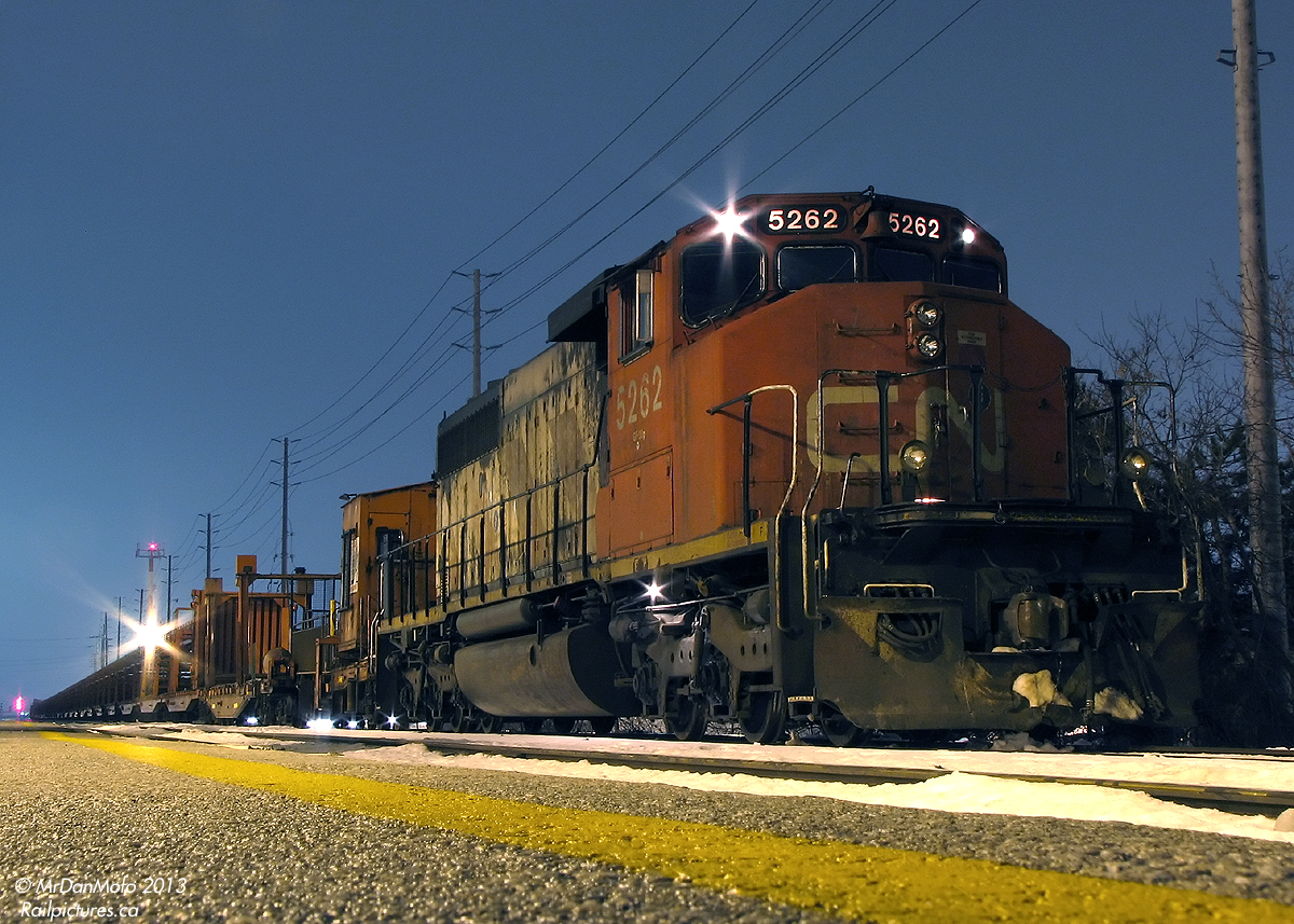 A Hard Day's Night

And after working like a dog all day laying rail along the Weston Sub, CN SD40-2W 5262 sits on point of the continuous welded rail (CWR) train, tied down for the night at the Malton GO Station. Sole power, the ratty 5262 has seen better days: its original 70's zebra stripe paint is now faded and rusty from many years of hard use. The CWR equipment behind it bears Transcona Shops (Winnipeg MB) logos, no doubt where it was built up from old flatcars by installing racks and winch equipment to facilitate picking up, storing, and laying long spaghetti-like strands of steel rail. In the distance, the eastbound signals at Woodbine West glow red, protecting the PM rush of westbound GO trains from any unauthorized track movements against them.

And the intrepid photographer, having gotten off the warm confines of the previous GO train for a shot or two, exposes a few more photos in the cold night before the next one home arrives.