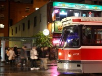 On a rainy night downtown, passengers waiting outside O'Gradys pub at McCaul rush onto the street to get aboard their eastbound College streetcar before getting wet. As is one of the rules of driving in downtown Toronto (and frequently elsewhere with streetcars) traffic in the curb lane must yield at designated stops to passengers boarding, in this case CLRV 4137, in the middle of the roadway.