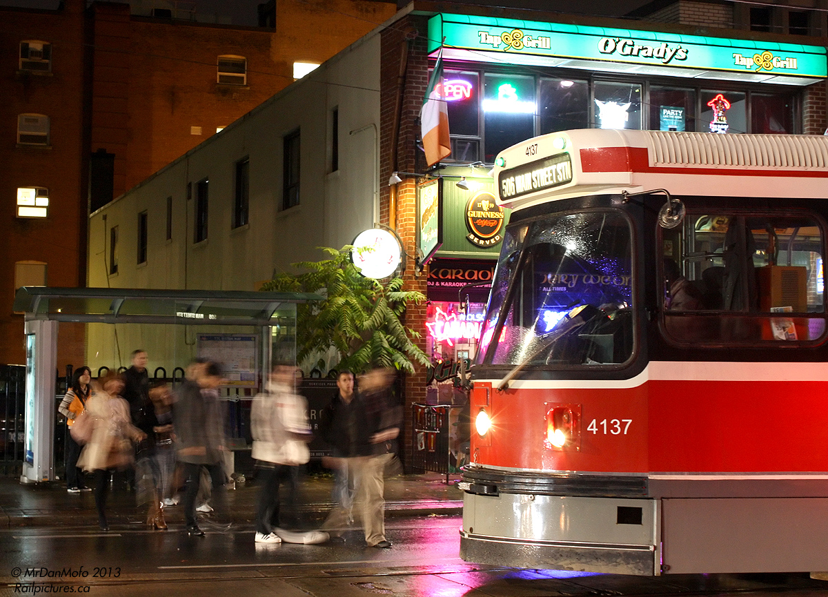 On a rainy night downtown, passengers waiting outside O'Gradys pub at McCaul rush onto the street to get aboard their eastbound College streetcar before getting wet. As is one of the rules of driving in downtown Toronto (and frequently elsewhere with streetcars) traffic in the curb lane must yield at designated stops to passengers boarding, in this case CLRV 4137, in the middle of the roadway.