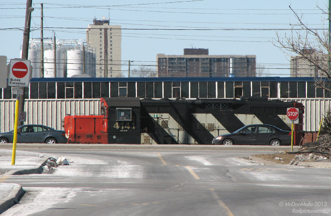 "Locomotive in Traffic"Suburban Brampton drivers on Steeles Avenue get momentarily distracted as CN 4141 rumbles by the busy thoroughfare with local 578. After finishing switching the industries in the Torbram/Queen/Steeles/Airport Rd. area, 578 is heading back to the mainline to tie onto their train on a sunny winter's afternoon. Extreme telephoto shot taken from near the Bramalea GO Station bus loop area (note the "hills" in the road), making for an interesting perspective.