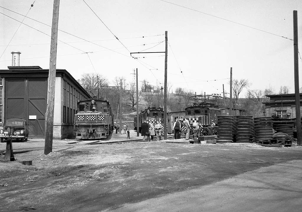Grand River Railway's Preston Shops is shown here full of freight and passenger equipment. GRR motor 230 is shown beside the shop building on the left, its end banner checker boards were installed in fall of 1949 and removed in 1953 when the motor was overhauled and rebuilt, helping to confirm the photo was taken in 1950. Beside it on the right is LE&N motor 333, and beside that is one of the three “big” motors, LE&N motor 337. Motor 337 was built by Preston Car and Coach in 1921 with two sisters, GRR 226, 228 as shells. The finishing electrics, all from Westinghouse, were installed by the Preston GRR shops. In the mid 1950s these motors (as were 9 of the 10 motors used on the railway) were rebuilt, made much heavier, and some were given larger traction motors.  On the right are piles of wheels. Preston had their own truing machine in the shop and in 1949-1950 replaced all of the wheels and tires on all of the passenger cars with larger diameter wheels. These wheels piles are part of that exercise.  In 1961 the line was dieselized by parent CPR using SW1200RS units, which were based out of Preston Shops. Today all that remains in this scene is the power house at right, and the mainline track (behind the photographer, and curving in the distance in the background far behind the engine building in photo above).  Photo taken by Cecil Hommerding, from the collection (Copyright) of Doug Leffler. Substantial caption information provided by George Roth et al with much thanks.  Other photos at Preston: GRR freight motor 230 at Preston Shops: http://www.railpictures.ca/?attachment_id=10297 LE&N freight motor 333 at Preston Shops: http://www.railpictures.ca/?attachment_id=10747 GRR passenger motor 848 at Preston: http://www.railpictures.ca/?attachment_id=10298  For more details on Cecil, see here.