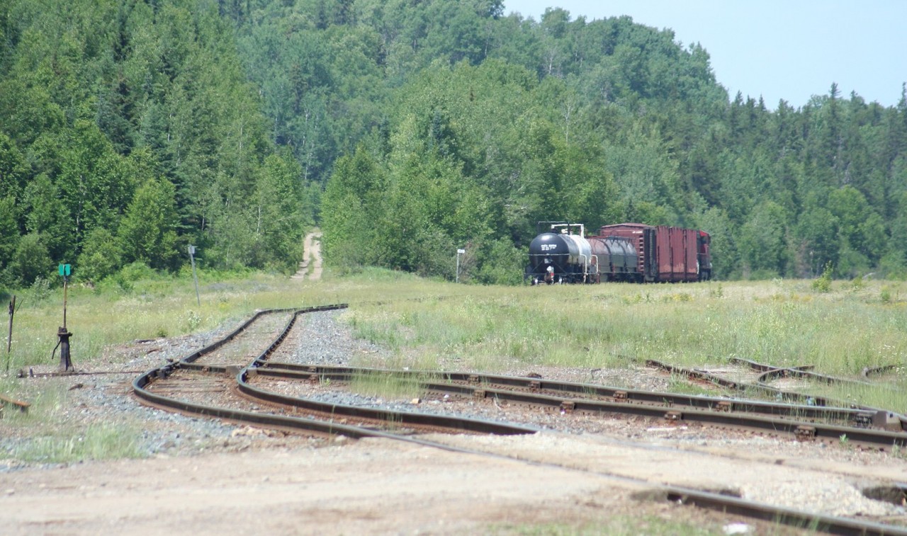 A short 9-car CN 571 trundles northward out of the desolation of the former Hawk Junction yard. The dirt track leading uphill in the background is the Algoma Central's former Michipicoten subdivision to Wawa and Michipicoten harbour, which was officially abandoned and the tracks lifted in 2000. The titular junction at Hawk Junction was once located just past the single remaining switch at left foreground leading to the south yard, which once commonly received trains of processed iron ore from the branch, but on this day saw only a lonely pulpwood flatcar. The grassy expanse to the right was once the main yard at Hawk Junction, where freight trains were made up and cars sorted. Now most of those tracks are completely ripped out, and the area becoming overgrown with grasses and weeds. (The track that disappears in the long grass in the centre of the image was formerly the ladded track for this part of the yard; you'll note a distinct lack of switchstands and points on this track today.) 


In it's heydey, Hawk Junction was a division point and the hub of operations on the Algoma Central Railway, seeing two daily regularly scheduled freights plus as required extras between Hawk Junction and Steelton yard in Sault Ste. Marie (in each direction), a daily freight between Hawk Junction and Hearst (again each way) and daily/as required ore trains on the branch to Wawa and Michipicoten Harbour as well as daily passenger service along the line. Today, the yard is barely more than a passing siding and a couple of set-out tracks, and both passenger and freight traffic is down to a thrice-weekly service. That little 9 car local trundling sure is a far cry from what once was.