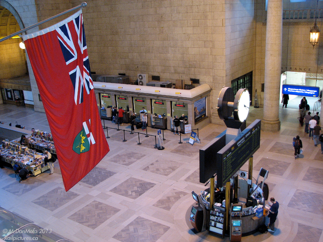 People linger in Toronto Union Station's Great Hall on a lazy Sunday afternoon. Some buying tickets from the customer service agents at the wickets, some browsing for a good book to read on the train before theirs is due to depart, others waiting for loved ones and friends to arrive on inbound trains from far off places across the province and country. The Flag of Ontario watches on over all, one of the many flags that adorn the walls of the Great Hall near the upper passageways of the station.