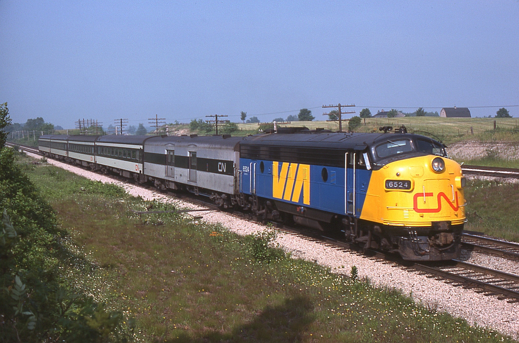 Motoring eastbound for its next stop at London, CN "VIA" train 84 with 6524 on point rolls through Hyde Park at Denfield Rd. The lead FP9 is in the early VIA paint with the CN noodle on the nose and the angled yellow partition under the cab, while the trailing 4 passenger cars are in the standard CN passenger fare of white and black. The baggage car appears to be the one specially painted black and silver to match the ex-Reading Crusader cars CN acquired (a photo of which can be seen here).