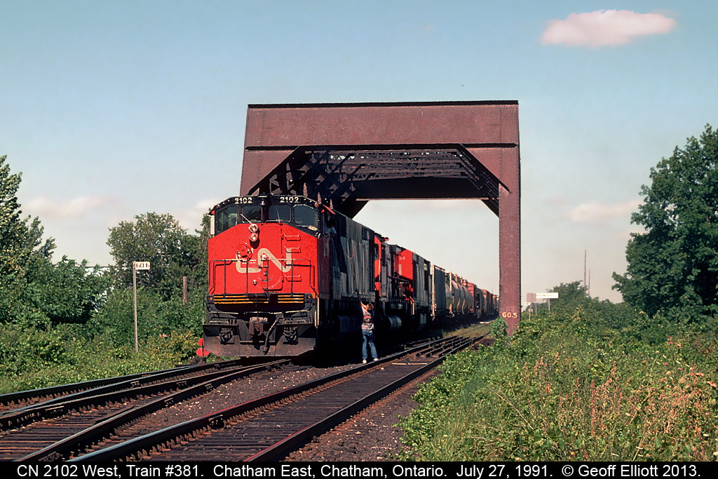 CN HR616 #2102 has train #381 down to a crawl as it readies to negotiate the cross-overs at Chatham East on July 27, 1991.  The Chatham East Operator has hoop and orders in hand as the Brakeman on 381 leans down to snag them.  2102 will go through the cross-overs and diverge onto the CSX Sarnia Sub for it's short run to Fargo, where the train will again turn west onto the CASO to finish out it's run to Windsor.