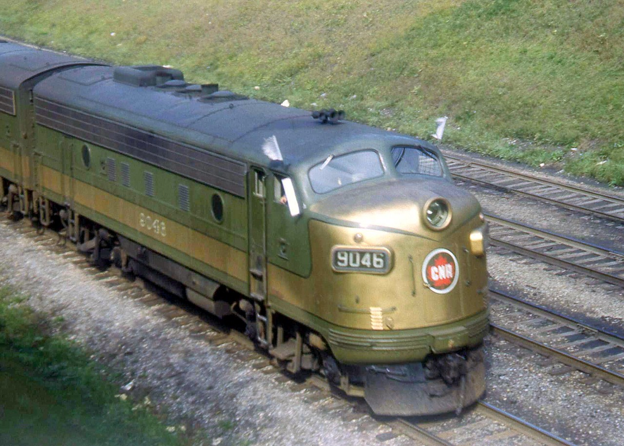 It's 1952 and CN F7A 9046, built July 1951, is nearly a year old as it heads eastbound through Sunnyside (Toronto) on the Oakville Sub, with white extra flags flapping in the wind.  The green & gold paint livery was the original delivery scheme for these units from GMD London, and was replaced with CN's yellow, green and black scheme in 1954, followed by the noodle and zebra stripes in 1961. Sunnyside Station and its platforms also still existed at this time, and the Gardiner Expressway that now parallels the Oakville Sub was still a few years away from construction. This image was taken from the south side of a now-gone footbridge that spanned the tracks between King St. and the Lake Shore Blvd., approximately half way between the station and Dowling Ave., likely removed when the Gardiner was built.   [Editor's note: accepted due to rarity]