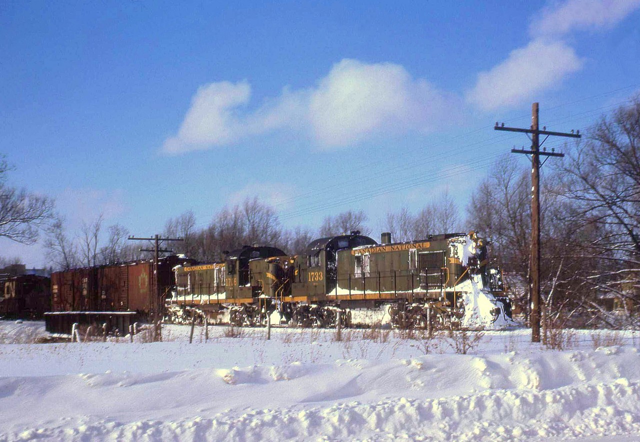 CN RSC-13 units 1733 and 1706 haul a freight train at Harriston ON, in January 1963. Harriston was Mile 5.01 on the CN's now-abandoned Owen Sound Subdivision (the line from Palmerston to Owen Sound), and junction point with the Southampton Sub (Harriston to Southampton). While the new CN noodle logo and image was introduced 2 years earlier as evident by the boxcar on the far left, the older green/yellow livery with the maple leaf herald still has a noticeable presence.  The RSC-13 was a CN-only model built by MLW in the mid-late 50's, with an RS3-style hood and A1A trucks but using the older 1000hp 539T engine - essentially their version of a GMD GMD-1. They were intended for light rail and branchline use, and while many worked out of Stratford and Toronto in their early years, they were more known for handling freight on Prince Edward Island in their later years (when retired, the A1A trucks from many went under RS18's upgraded for branchline service as RSC14's).  Geotagged location not exact.