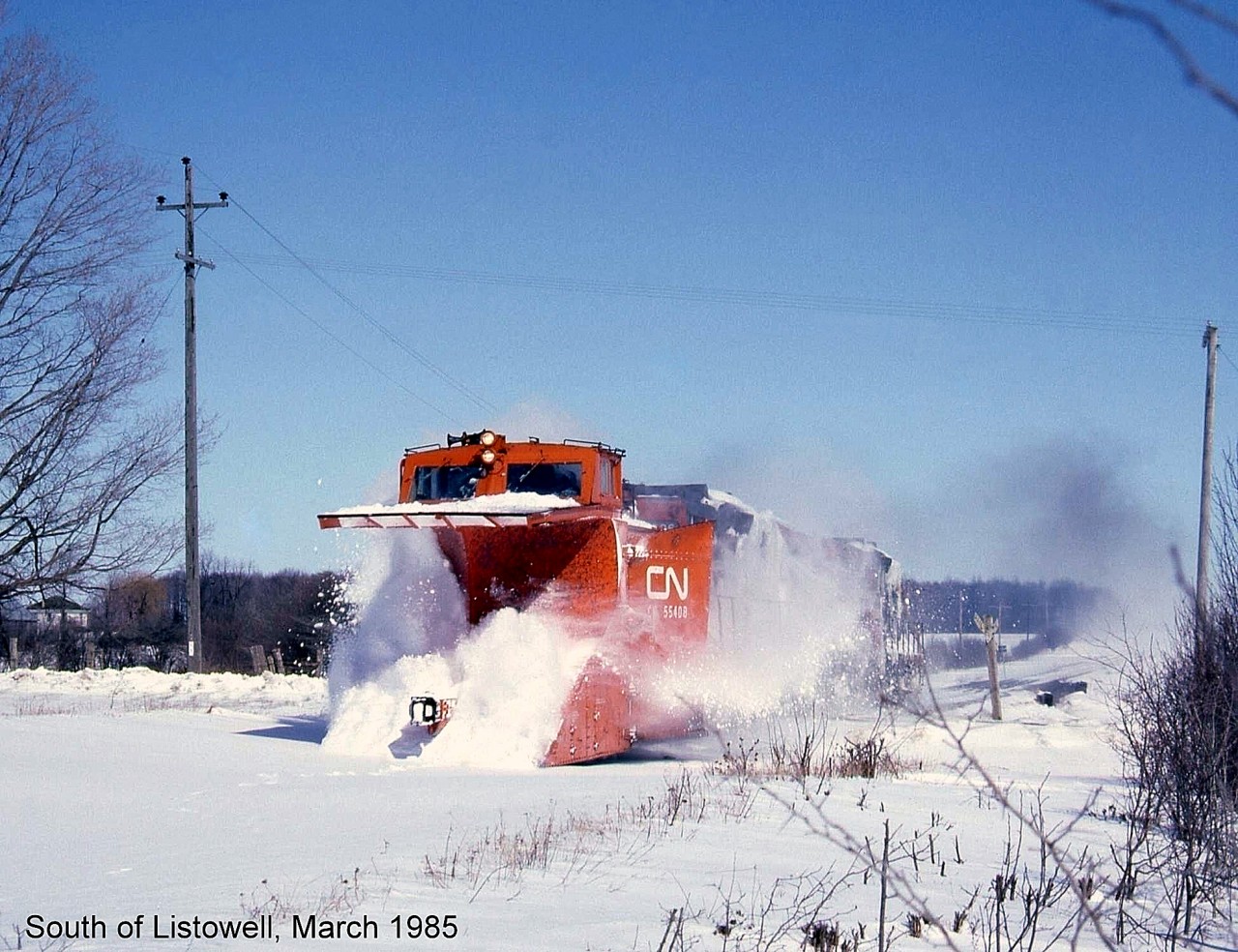 I was working in the Listowell area in March of 1985 and heard on my scanner that there was a plow extra heading south on the CN Newton Sub (later west) to Kincardine. I had time to go to a side road south of town and set up near a crossing and the plow soon appeared. I took my photo just before it passed then WAM, I was bowled over by a sheet of heavy snow and knocked over a fence (I was not injured but it was a shock). I then went to a local restaurant and tidied up my clothing. The resulting photo was average but it brings back memories: CN snowplow 55408 charging across a grade crossing south of Listowel, propelled by a pair of GP9 units.

[Editor's note: CN plow 55408, a wedge plow built by Eastern Car Co (Trenton NS) in 1937, mets its end not too long ago after avoiding the scrapper once. Once it was retired from CN in the 90's (many lines being abandoned or torn up in the southern Ontario area reduced the need for plows) it was sold to Zubicks in London for scrap. In 1995 the Goderich Exeter Railway traded their plow, 55437, to Zubicks for 55408, as it was in better shape. Known by some as their "orange plow" (they also owned sister 55413 painted black), it continued the fight on mother nature for its new owner until recently, when it was cut up for scrap in Goderich during May 2013.]

(Note, geotagged location may not be exact)