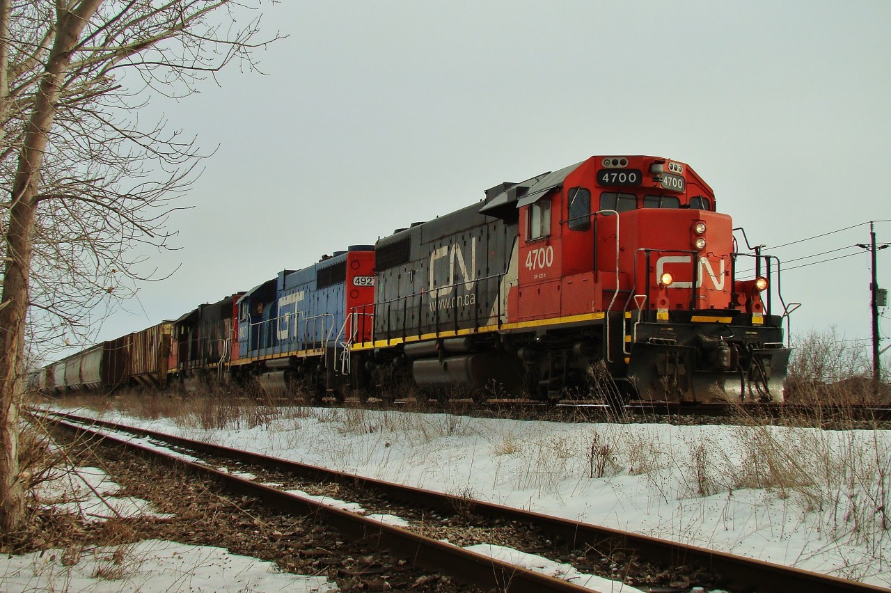 Looking through a hole in the fence, we see CN 439 heading up the grade to Dougall Ave. lead by CN 4700, GTW 4927 and CN 4710 with a 40 car train bound for Van de Water yard. The spur in the foreground heads to Zalev's Scrap.
