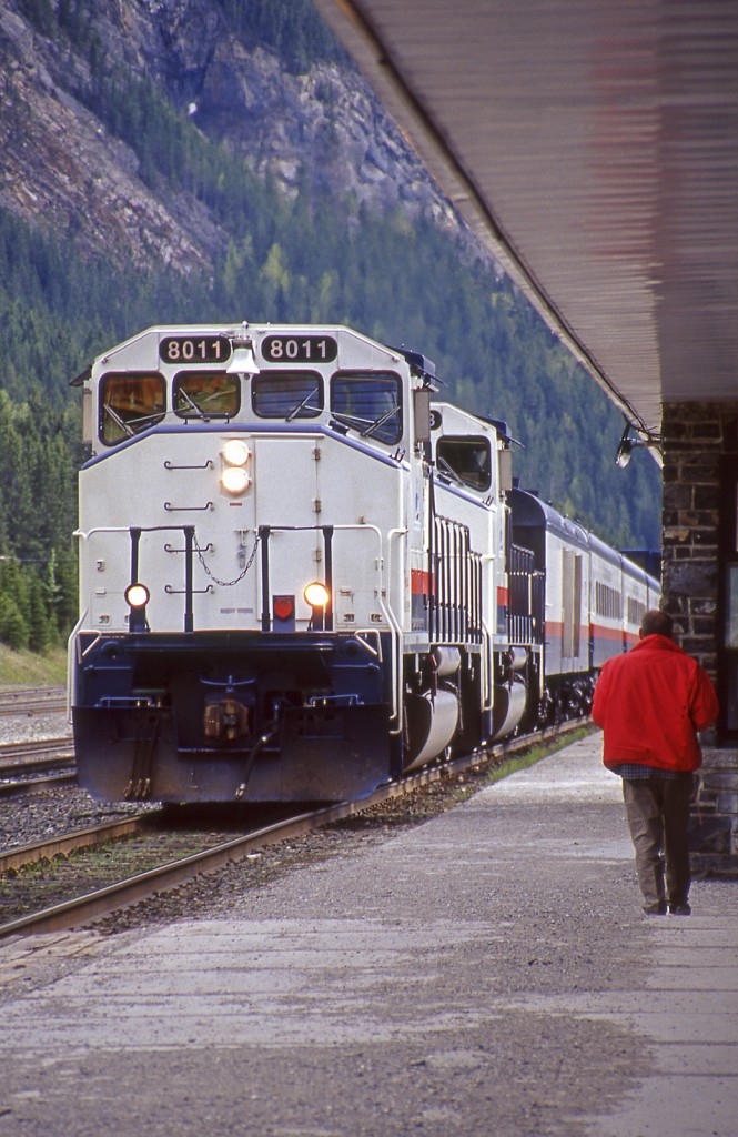 The eastbound Rocky Mountaineer Railtours passenger train rolls up to a station stop as a lone passenger (actually my father ) awaits to greet the train.