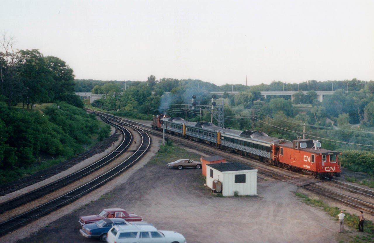 While waiting early one evening for a CN westbound, which was approaching just around the corner; I happened to see this oddity sneaking by, heading east. So what have we here? A guess would be a breakdown of the afternoon Niagara Falls Budds; resulting in the consist being towed back, complete with caboose. Bill McArthur and Dave Stowe (right) watch the "extra" accelerate thru the junction with CN 1264, Budds 6302, 6005 6120 and Van 79534. Jalopies in the foreground belong to Bill (blue wagon) Steven Lee (maroon valiant) and my '76 Cougar in the middle. The tan car, a Chevy Nova, it looks like, was often present watching trains on weekends but I never did put a name to the owner. As this view indicates, this was a pleasant tranquil location back in the day for trainwatching. Why did so many hang out here? Well, it was the days before technology as we now know it, and to find out the 'train-world happenings' one stopped by to get the latest, so the junction was the place to be.
