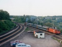 While waiting early one evening for a CN westbound, which was approaching just around the corner; I happened to see this oddity sneaking by, heading east. So what have we here? A guess would be a breakdown of the afternoon Niagara Falls Budds; resulting in the consist being towed back, complete with caboose. Bill McArthur and Dave Stowe (right) watch the "extra" accelerate thru the junction with CN 1264, Budds 6302, 6005 6120 and Van 79534. Jalopies in the foreground belong to Bill (blue wagon) Steven Lee (maroon valiant) and my '76 Cougar in the middle. The tan car, a Chevy Nova, it looks like, was often present watching trains on weekends but I never did put a name to the owner. As this view indicates, this was a pleasant tranquil location back in the day for trainwatching. Why did so many hang out here? Well, it was the days before technology as we now know it, and to find out the 'train-world happenings' one stopped by to get the latest, so the junction was the place to be.