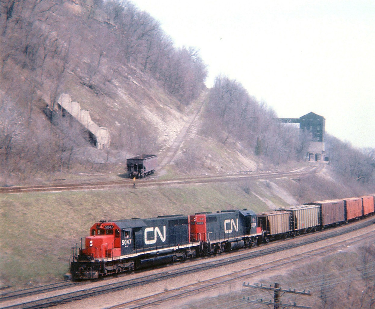 CN 5047, 5525 westbound pass the old Canada Crushed Stone Ltd facility near mile 4, Dundas Sub. This image, while a tad grainy, shows a rather open view of the old rail siding which led up to the large screen house . Operations before shutting down in the early 1970s consisted of stone quarried up on the mountain brow (Dundas Peak)going thru the High Brow Crusher (demo'd 1975) and then down to the Screen House (demo'd 1993)and then over the CN mainline via the gantry (demo'd 1986)conveyer belt and stockpiled on the south side for later shipping. Interesting in this image is the hopper at the base of the incline trackage. This steep grade operated by cable pulling cars up the grade, supplies usually went this way. Perhaps this car was held back for use in removal of some equipment as the shutdown was finalized. Steetley Inc., which purchased the CCSL in 1951, maintained a small shipping facility south of the tracks until the early 1980s. Walking the area today, it is hard to imagine the landscape being so open. All that remains now is the incline bed, which is now a hiking trail; all rail is gone but a lot of old ties remain.