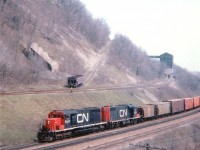 [Editors note: historic] CN 5047, 5525 westbound pass the old Canada Crushed Stone Ltd facility near mile 4, Dundas Sub. This image, while a tad grainy, shows a rather open view of the old rail siding which led up to the large screen house . Operations before shutting down in the early 1970s consisted of stone quarried up on the mountain brow (Dundas Peak)going thru the High Brow Crusher (demo'd 1975) and then down to the Screen House (demo'd 1993)and then over the CN mainline via the gantry (demo'd 1986)conveyer belt and stockpiled on the south side for later shipping. Interesting in this image is the hopper at the base of the incline trackage. This steep grade operated by cable pulling cars up the grade, supplies usually went this way. Perhaps this car was held back for use in removal of some equipment as the shutdown was finalized. Steetley Inc., which purchased the CCSL in 1951, maintained a small shipping facility south of the tracks until the early 1980s. Walking the area today, it is hard to imagine the landscape being so open. All that remains now is the incline bed, which is now a hiking trail; all rail is gone but a lot of old ties remain.